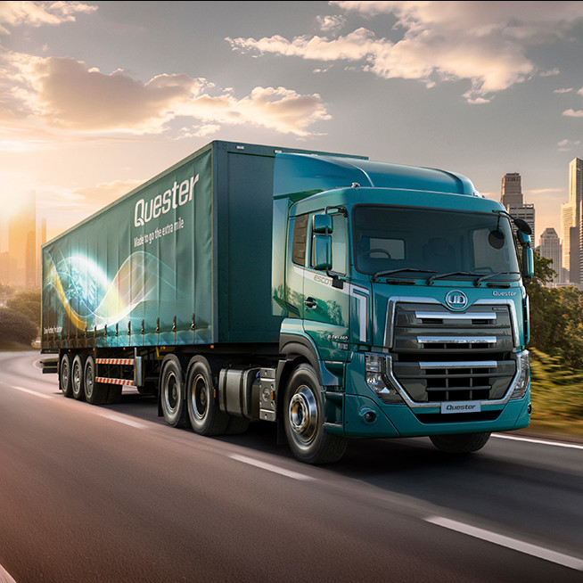 Blue semi-truck drives on highway toward city skyline at sunset, trees line the road.