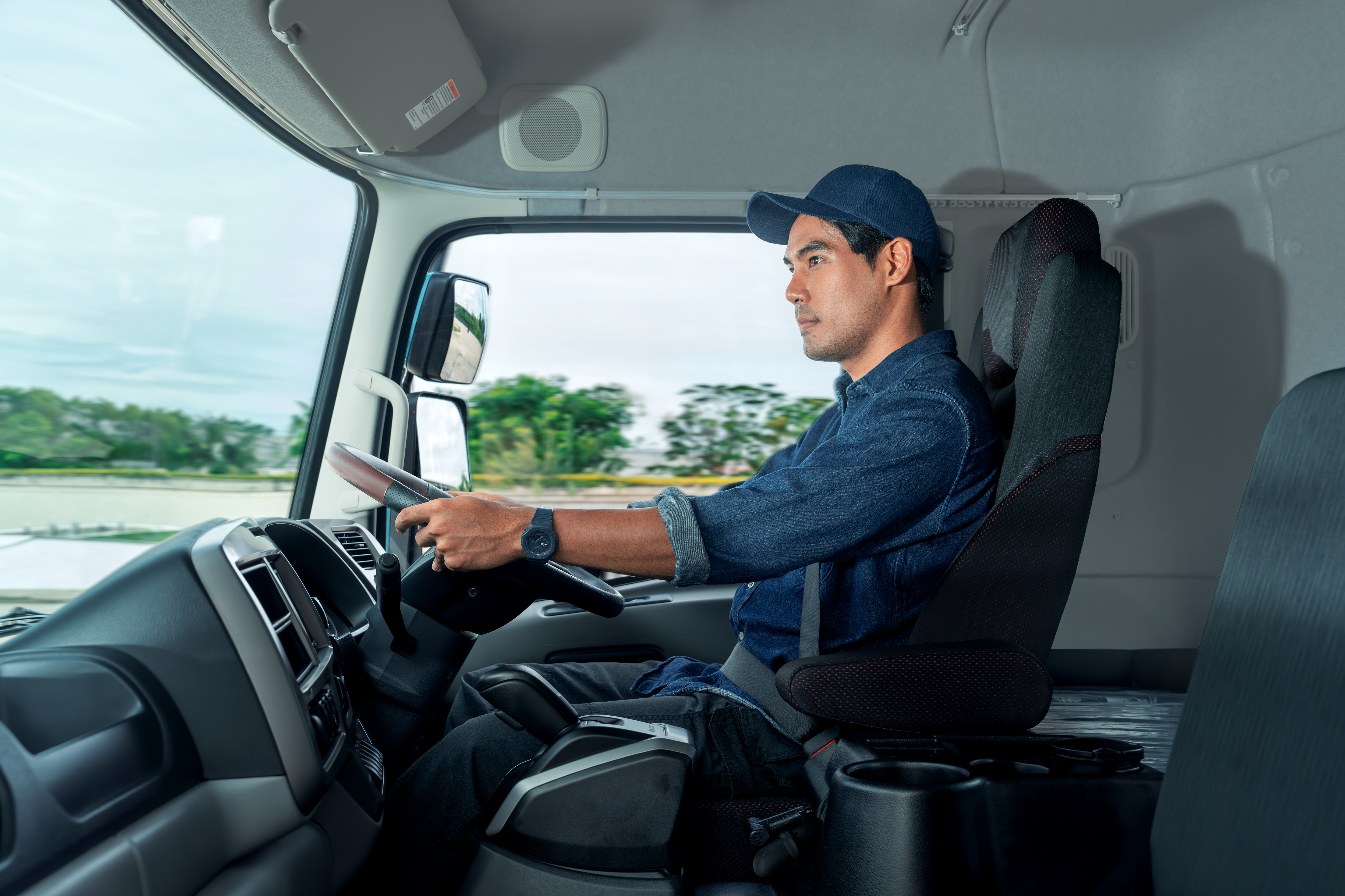 Quester Man in blue uniform driving a truck, focused on the road, trees visible outside window.