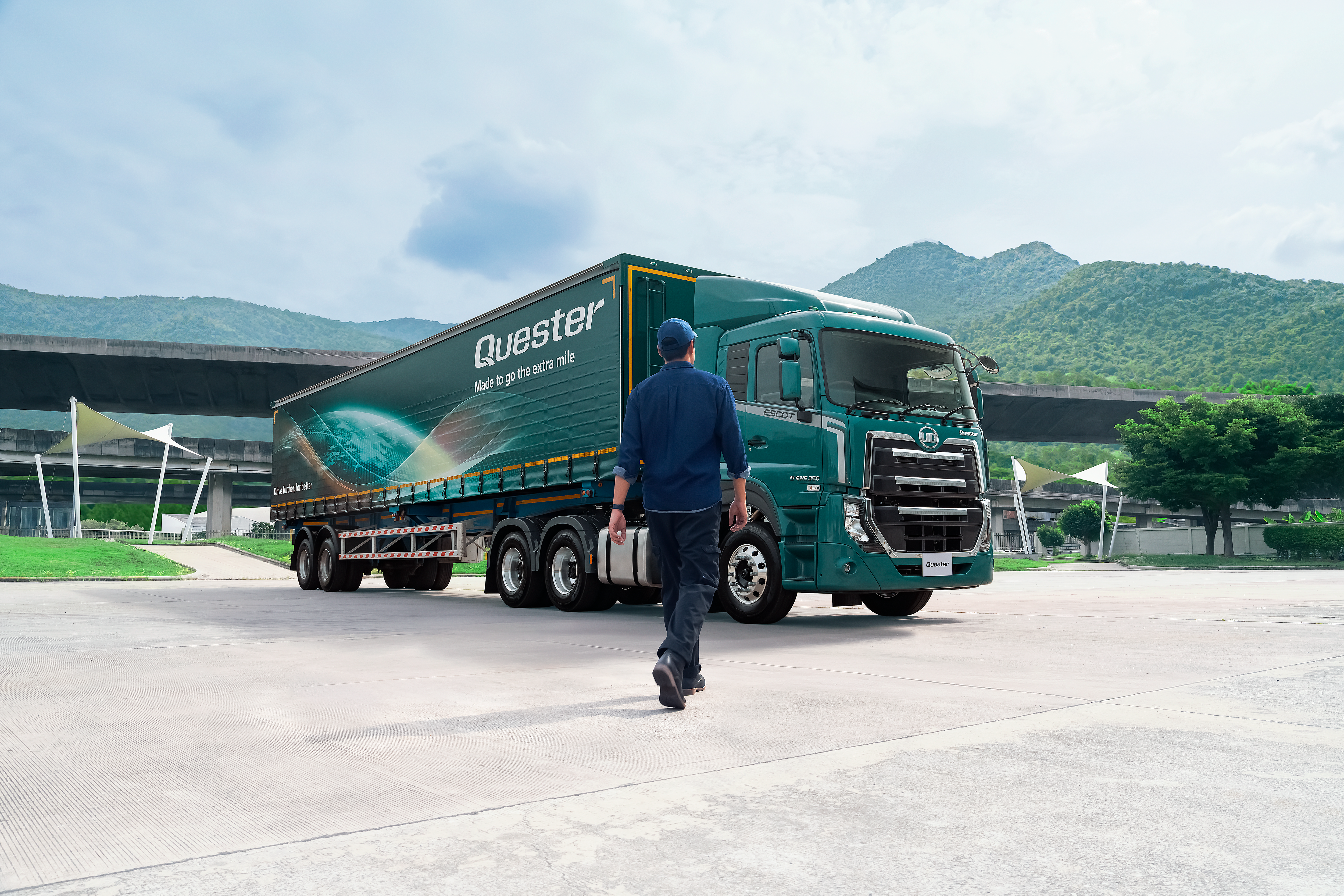 Quester Person walking toward a teal semi-truck in an outdoor lot with mountains in the background.