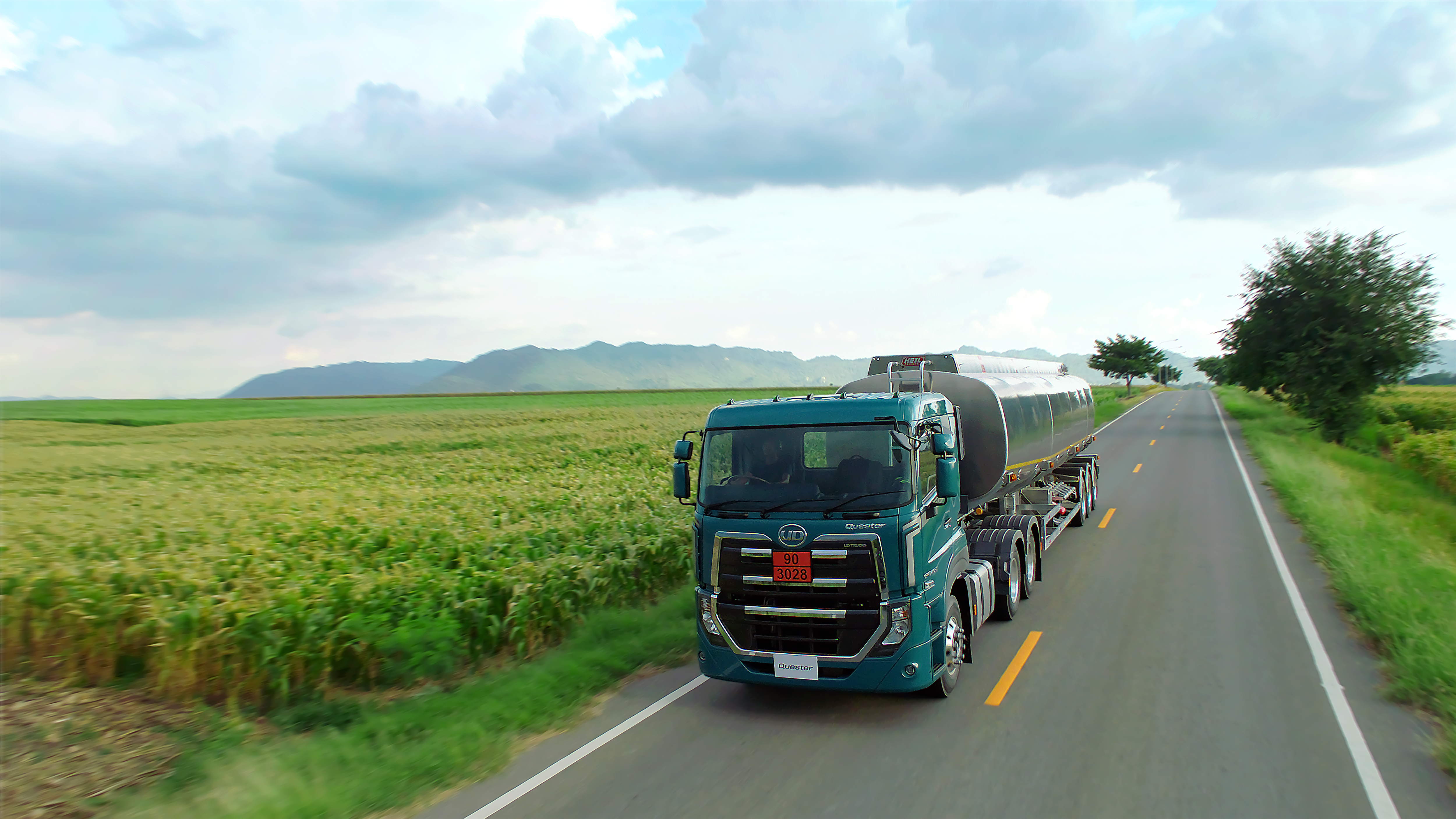Quester Tanker truck driving on rural road between green fields under a cloudy sky.