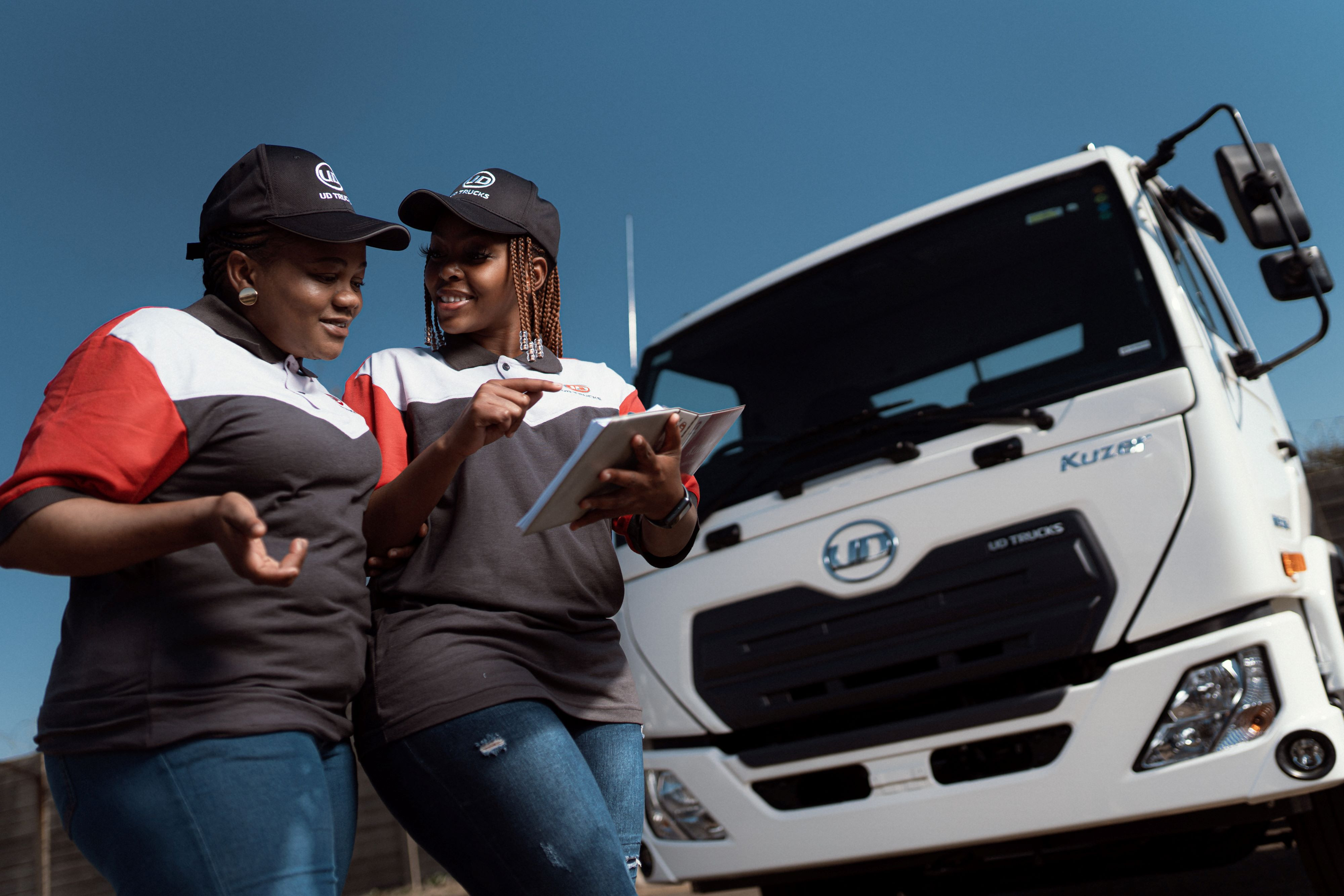 Twee vroue gesels en hou ’n tablet langs ’n wit vragmotor in sonskyn.