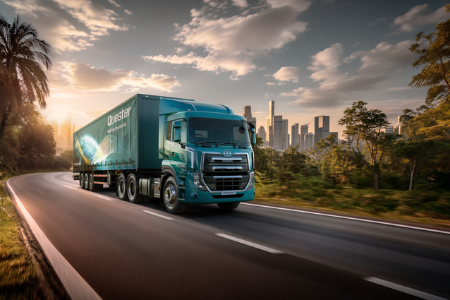 Blue semi-truck driving on a road with a city skyline in the background at sunset.