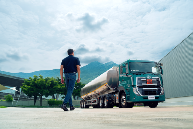 Man walks toward a green semi truck on a sunny day near mountains and industrial buildings.