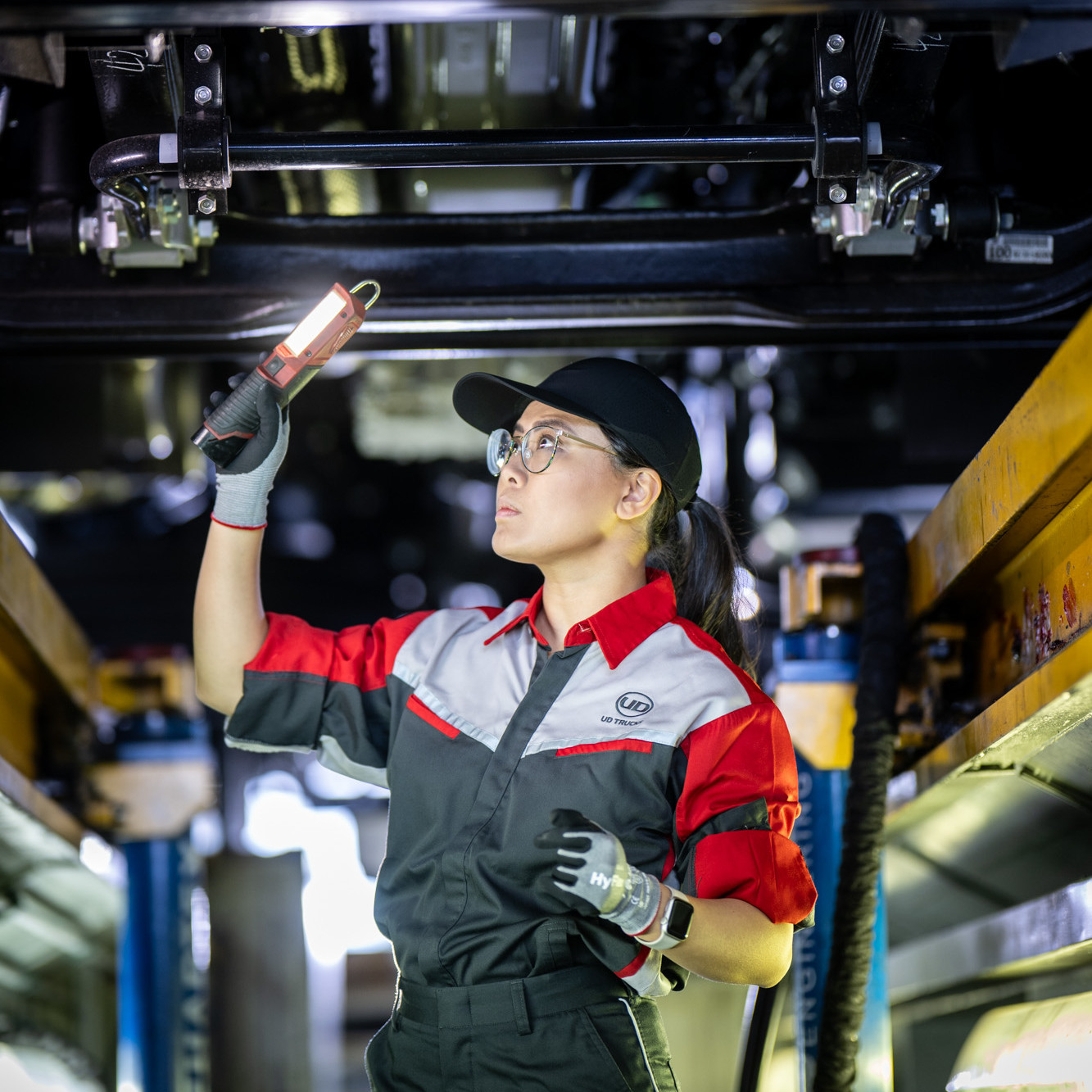 Mechanic in uniform inspects under a vehicle with a torch in a workshop.