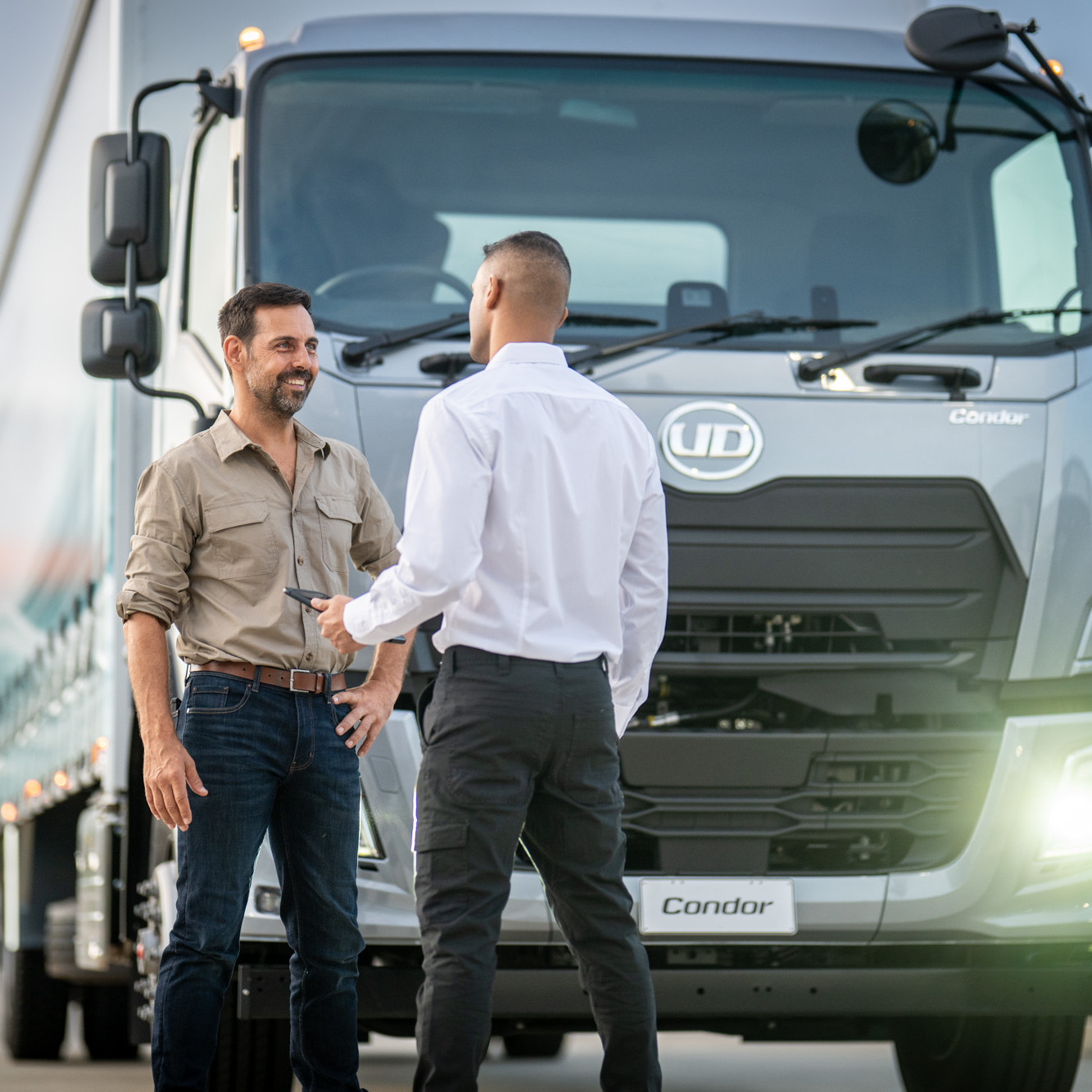 Two men talking in front of a large silver truck in daylight.
