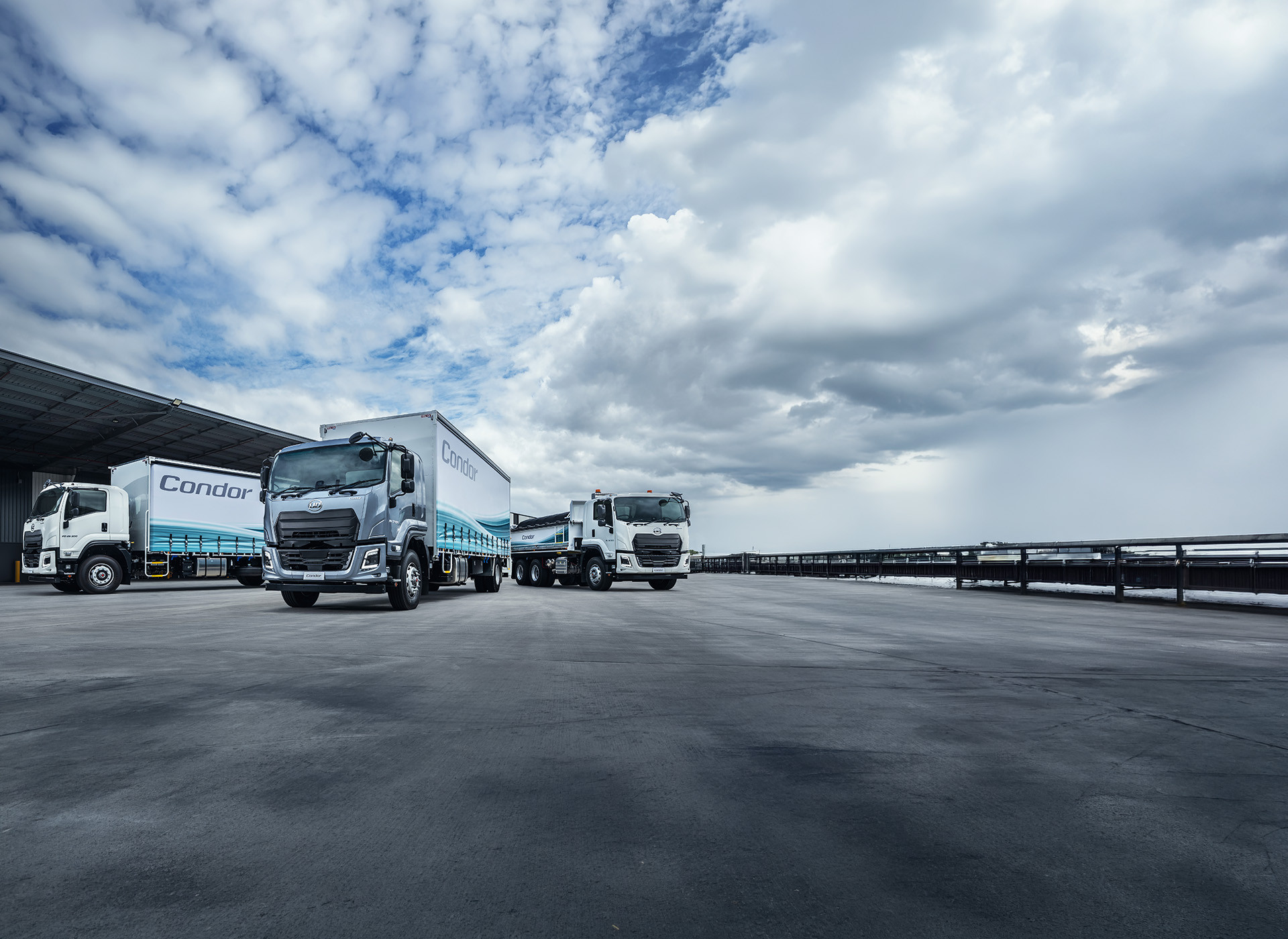 Three large delivery trucks parked beside a warehouse under a cloudy sky.
