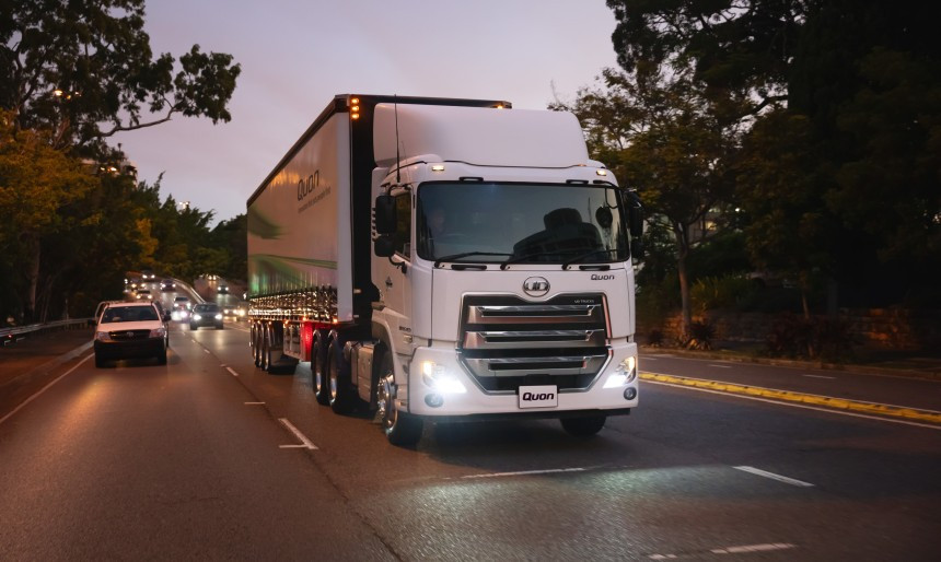 White semi-truck driving on a road at dusk, headlights on, surrounded by trees and cars.