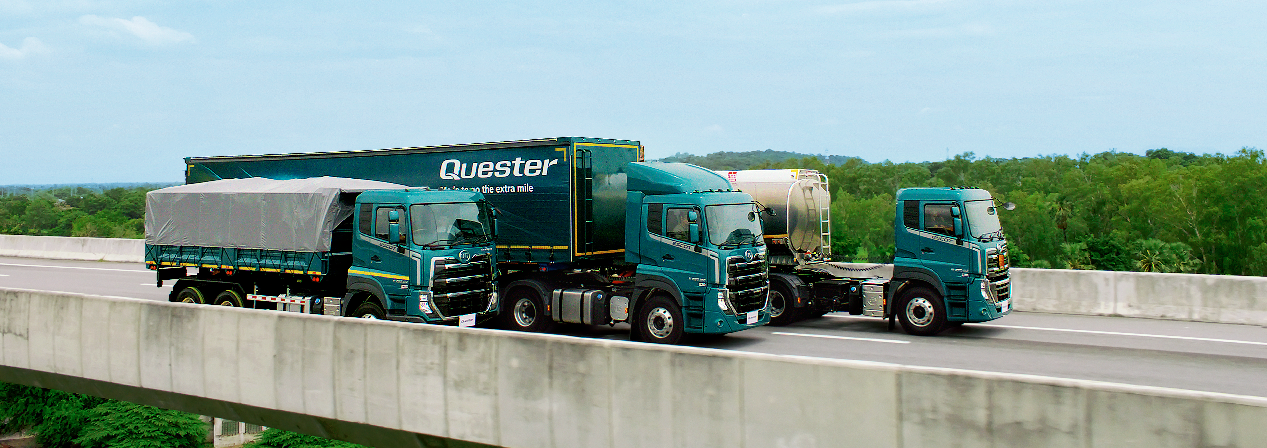 Three teal Quester trucks on a bridge with green trees in the background under a blue sky.