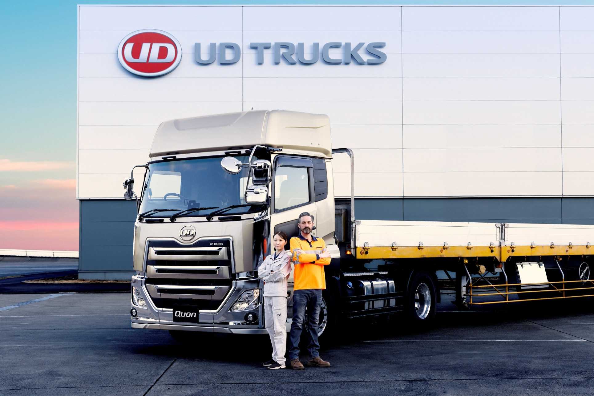 Two people standing beside a large silver truck outside a UD Trucks building at sunset.