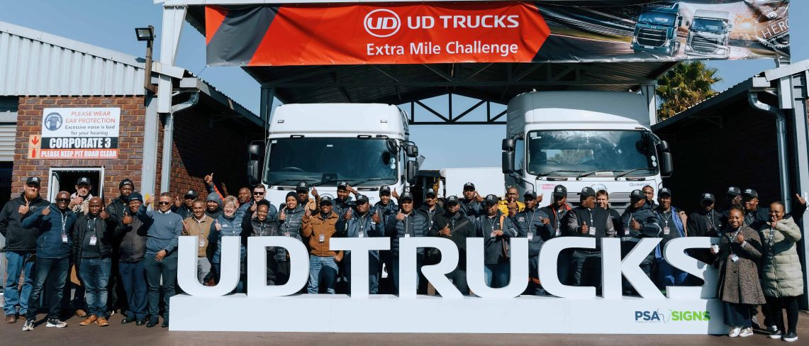 Groot groep mense poseer voor twee wit vragmotors by 'n UD Trucks-byeenkoms.