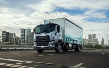 Large silver delivery truck driving on a city road with modern buildings in background.