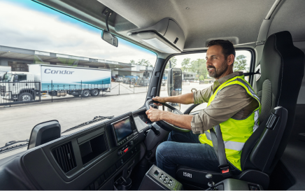 Man in high-vis vest driving a truck near a warehouse and parked lorry.
