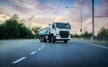 White semi-truck driving on a curved road at sunset, trees in background.