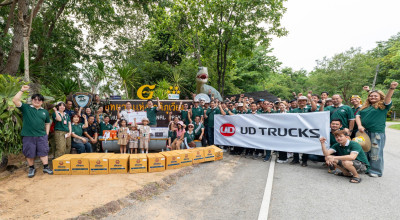 กลุ่มคนยืนถ่ายรูปกลางแจ้งถือป้าย UD TRUCKS ท่ามกลางต้นไม้และธรรมชาติ