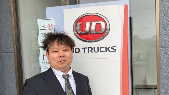 スーツ姿の男性がUD TRUCKSのロゴ看板の前に立っている