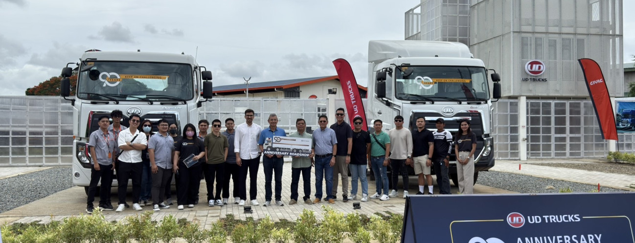 Grupo ng tao sa harap ng dalawang trak, may gusali at event banner sa likod.