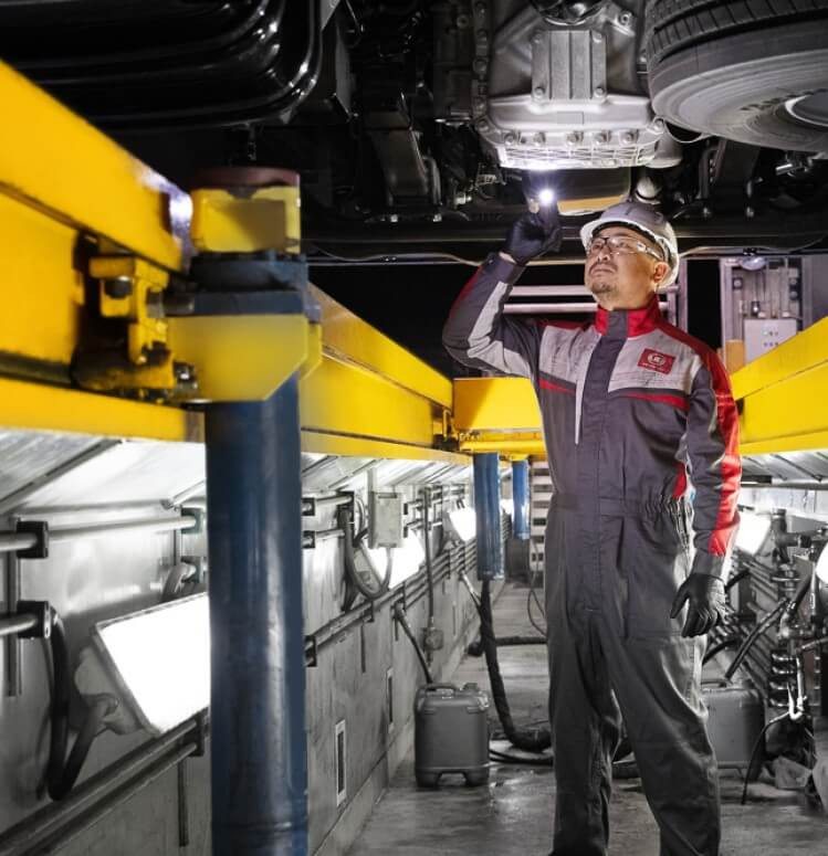 Mechanic in uniform inspecting underside of a vehicle with a flashlight in a workshop.